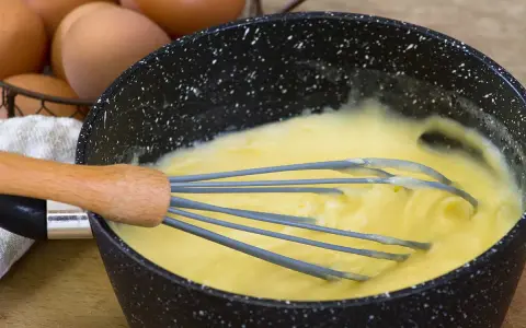 Un fouet mélange une crème pâtissière traditionnelle onctueuse dans une casserole. Des œufs sont visibles en arrière-plan.