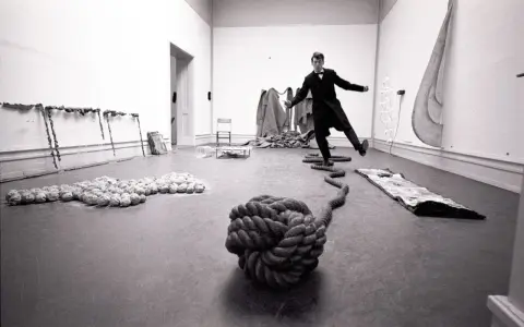 Un homme en costume, peut-être Lawrence Weiner, danse au milieu d'une installation artistique avec des cordes et des objets.