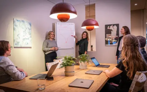 Ett team diskuterar strategier vid ett konferensbord med flera laptops. En kvinna pekar på en trattmodell på en whiteboard. Ups datorn är en del av deras effektiva arbetsflöde.