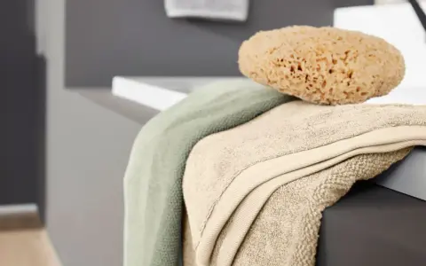 Weiche bad textilien in sanften Farben, darunter ein hellgrünes und ein beigefarbenes Handtuch, liegen neben einem natürlichen Schwamm auf der Kante einer Badewanne.