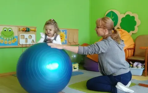 Dziecko w okularach ćwiczy z fizjoterapeutką na dużej, niebieskiej piłce w sali przedszkolnej.