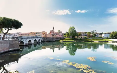 Ponte di Tiberio e il fiume Marecchia a Rimini a settembre, con riflessi sull'acqua e un cielo azzurro con nuvole bianche.