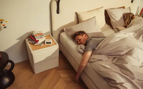 Mann schläft tief und fest auf einem Bett, das sich wie **Kaltschaum** anfühlt. Neben dem Bett liegen Bücher und ein Wecker.