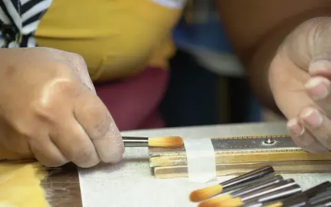 Une main tient un pinceau, prêt à peindre. D'autres pinceaux sont alignés à côté d'une règle.