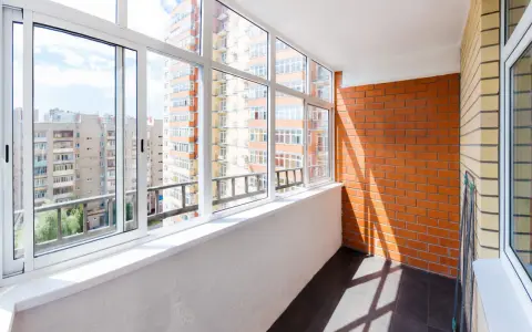 Wnętrze loggii z widokiem na miejską zabudowę. Ściany z cegły i tynku, przeszklony balkon z białymi ramami okiennymi.