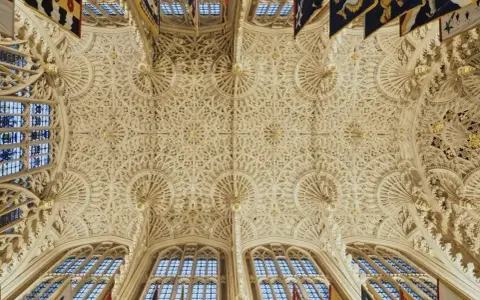 Intricate fan vaulting of a grand hall, reminiscent of the detailed ceilings found in abbey graves, adorned with flags.