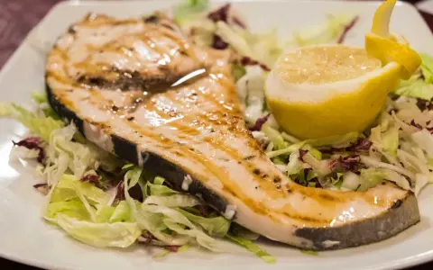 Pez espada a la plancha con ajo y perejil, servido sobre una cama de lechugas y acompañado de un gajo de limón.