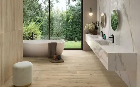 Salle de bain moderne avec baignoire îlot, double vasque en marbre et **pose carrelage imitation parquet** au sol et sur un mur. Vue sur jardin luxuriant.