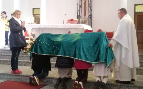 Dzieci klęczą pod zielonym welonem, odmawiając modlitwę do Matki Bożej z Guadalupe. Kapłan czuwa obok.