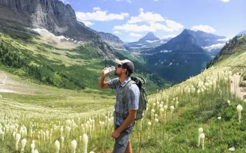 Randonneur buvant de l'eau au milieu d'un champ de fleurs blanches, avec des montagnes majestueuses en arrière-plan. Il espère que cela soulagera sa crampe cuisse.
