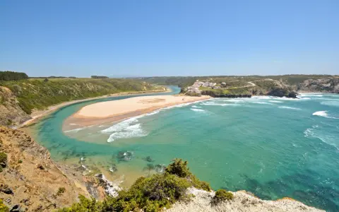 Atemberaubende Aussicht auf Praia de Odeceixe, wo ein Fluss auf den Ozean trifft. Grüne Hügel und ein weißes Dorf säumen die Küste.