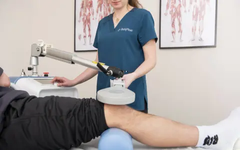 Fizjoterapeuta wykonuje zabieg rehabilitacyjny na kolanie pacjenta za pomocą specjalistycznego urządzenia.