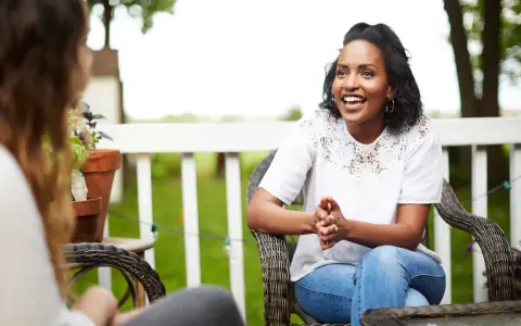 Two women are sitting on a porch, one animatedly sharing the gospel with the other.