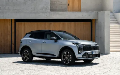 Elegancki, srebrny Kia Sportage Hybrid z czarnym dachem zaparkowany przed nowoczesnym budynkiem z drewnianymi drzwiami i betonowymi schodami.