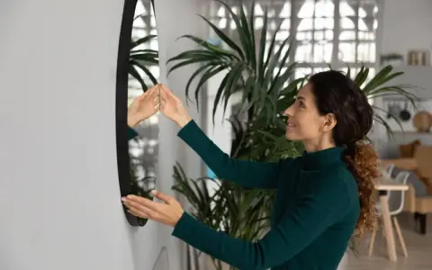 Mujer sonriente ajusta un espejo sin marco en la pared, mostrando cómo colgar un espejo sin marco para decorar su hogar.