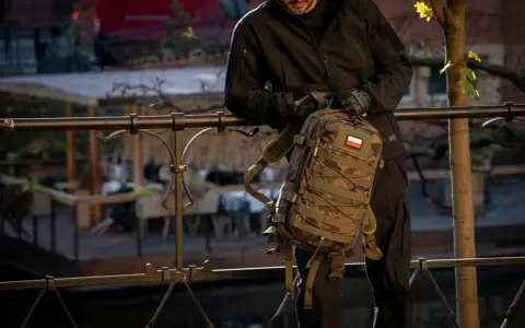 Mężczyzna w czarnej kurtce i rękawiczkach trzyma plecak w kamuflażu, z polską flagą. To jeden z wielu stylowych modeli marki plecaków.