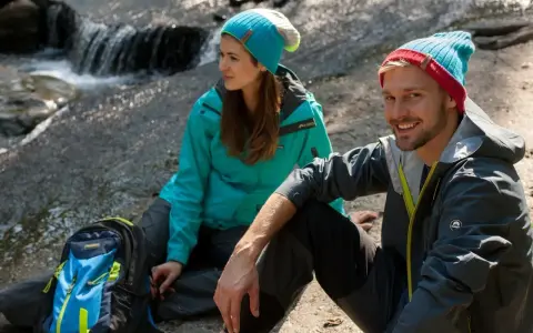 Para, co to jest trekking? Para w czapkach i kurtkach siedzi przy strumieniu, obok plecak.