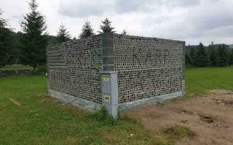 Nietypowy domek z butelek plastikowych, zbudowany z tysięcy szklanych opakowań, otoczony zielenią i drzewami.