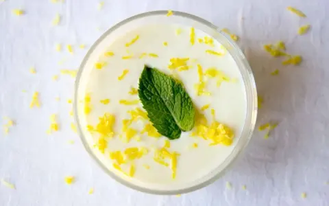 Delicioso mousse de limón, adornado con ralladura de limón y una hoja de menta fresca, servido en un vaso transparente.