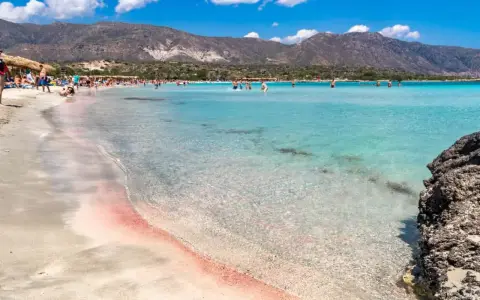 Spiaggia rosa di Creta con bambini che giocano nell'acqua cristallina e montagne sullo sfondo.