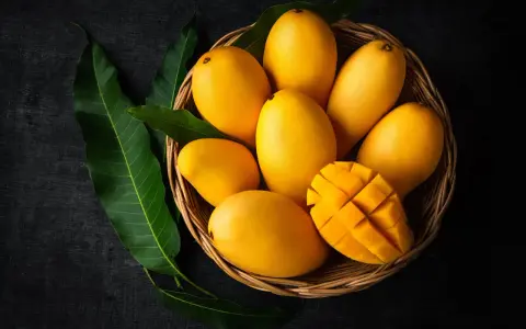 Koszyk pełen soczystych, żółtych mango, jedno przekrojone w kostkę. Obok zielone liście mango.