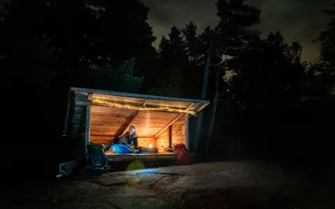 Person myser i ett upplyst vindskydd i skogen på natten. Ryggsäckar står utanför.
