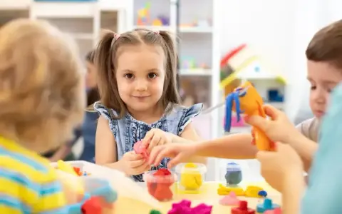 Uśmiechnięta dziewczynka z kitkami bawi się plasteliną, zastanawiając się, w jakim wieku do szkoły pójdzie. Dzieci tworzą kolorowe figurki.
