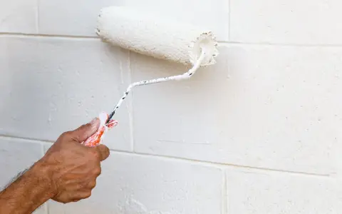 Main tenant, avis peinture carrelage sol : un rouleau blanc applique une nouvelle couche sur un mur en parpaings.