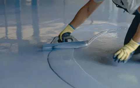 Persona con guantes amarillos aplica una capa de pintura azul clara sobre un suelo de terrazo con una llana.