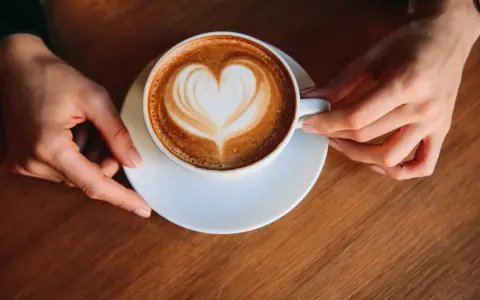 Eine Tasse Milchkaffee mit Herz-Latte-Art, gehalten von zwei Händen. Ein perfekter Start in den Tag, was ist Milchkaffee?