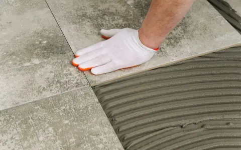 Ręka w białej rękawiczce układa płytkę gresową na kleju. Różnica między gres a płytkami jest widoczna w procesie układania.