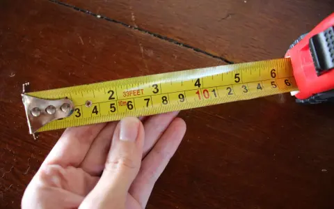 Una mano sujeta una cinta métrica amarilla sobre una mesa de madera. La cinta muestra medidas en pies y metros, demostrando para que sirve la cinta métrica: medir distancias.