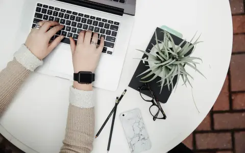 Dłonie piszą na laptopie, obok leżą okulary, telefon i roślina. Może to być błąd interfejsu, gdy coś nie działa.