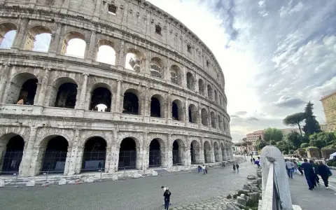 Le Colisée, monument emblématique de Rome, se dresse majestueusement sous un ciel nuageux. Des visiteurs explorent ses environs pavés.
