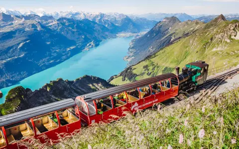 The Brienzer Rothorn train ascends a steep mountain track, offering passengers breathtaking views of a turquoise lake and snow-capped peaks.
