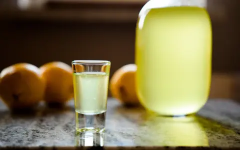 Szklaneczka domowego limoncello, obok cytryny i dużej butelki. Idealny przepis na orzeźwiający trunek.