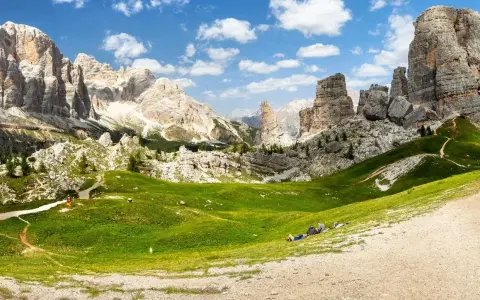 Un paesaggio mozzafiato nelle Dolomiti con le iconiche 5 torri che si ergono maestose. Un prato verde brillante invita al relax, con persone che riposano sotto il cielo azzurro.