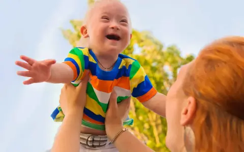 Niño con síndrome de Down, sonriente, es levantado por una persona. Sus rasgos son visibles y llenos de alegría.