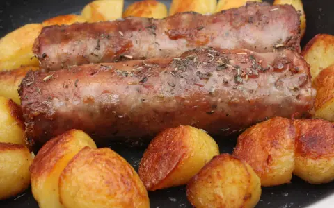 Deux andouillettes artisanales dorées, parsemées d'herbes, mijotent avec des pommes de terre rôties dans une poêle.