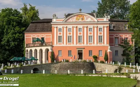 Kurozwęki pałac, z pomarańczową fasadą i białymi zdobieniami, otoczony zielenią. Na pierwszym planie mostek i trawnik.