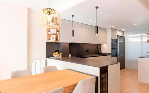 Moderna cocina en L con isla, electrodomésticos integrados, encimera de cuarzo y mesa de madera.