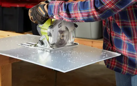 A person in a plaid shirt and gloves uses a circular saw to cut a sheet of plexiglass, creating small plastic shavings. This demonstrates that plexiglass is easy to cut with the right tools.