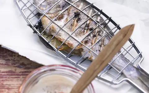 Ein ganzer Fisch liegt in einem Grillkorb, bereit zum Fisch grillen auf dem Elektrogrill. Kräuter und Zitronenscheiben sind im Fisch.