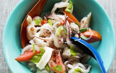 Ensalada tailandesa con fideos de cristal, calamares, tomate y cebolla, servida en un cuenco azul. El plato de sepia en salsa se ve fresco y apetitoso.