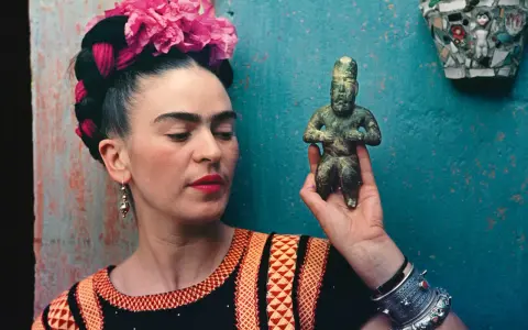 Frida Kahlo, avec ses fleurs roses et sa robe colorée, tient une statuette ancienne.