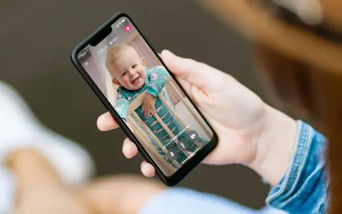 Ein lächelndes Baby im Strampler schaut aus dem Gitterbett. Eine Hand hält das Handy als Babyphone, um das Kind im Blick zu behalten.