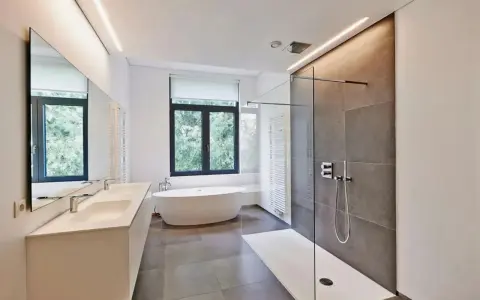 Salle de bain moderne avec baignoire îlot, double vasque et douche à l'italienne. L'éclairage intégré en hauteur applique une ambiance douce.