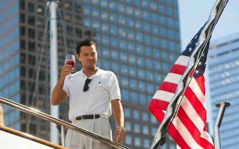 Leonardo DiCaprio z kieliszkiem wina na jachcie, obok amerykańska flaga i wieżowce.