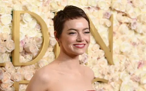 Emma Stone sorride, con un trucco che esalta i colori che stanno bene alle castane con pelle chiara. Lo sfondo è un muro di rose rosa e bianche.