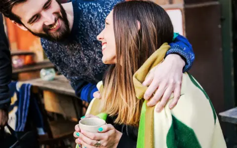 Pareja sonriente compartiendo una manta y una taza de té. Él le abraza por detrás. Ella le dice "muchas gracias en francés".